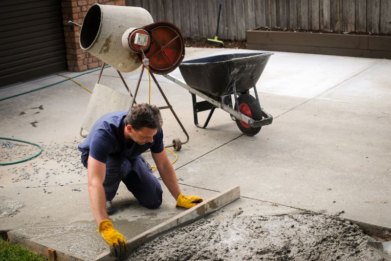 Tradesperson smoothing a fresh concrete patch beside a cement mixer and wheelbarrow